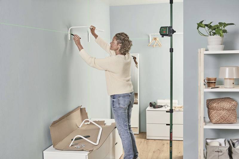 Eine Person montiert ein Regal an einer blauen Wand. Ein Laser-Niveau zeigt die gerade Linie. Im Hintergrund sind Werkzeuge und Möbel.