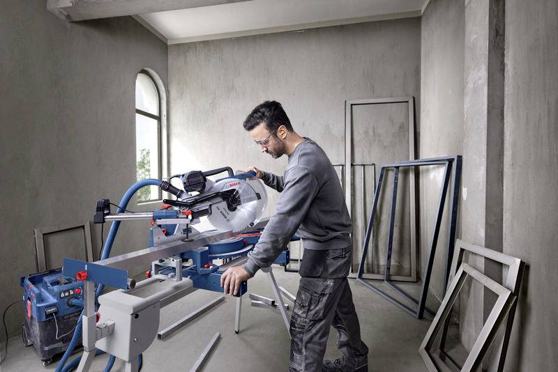 Ein Mann in Arbeitskleidung bedient eine Kappsäge in einer Baustelle. Im Hintergrund sind leere Fensterrahmen erkennbar.