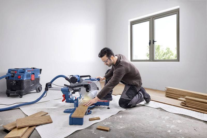 Mann schneidet Holzbrett mit einer Kappsäge in einem Raum. Staubsauger angeschlossen, Holzstücke gestapelt. Fenster im Hintergrund.