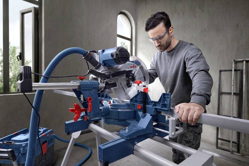 Ein Mann arbeitet in einer Werkstatt mit einer elektrischen Gehrungssäge. Er trägt Schutzbrille und konzentriert sich auf das Sägen.