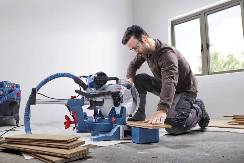 Ein Mann kniet auf dem Boden und schneidet Holz mit einer elektrischen Tischsäge in einem hellen Raum. Er trägt Schutzbrille.