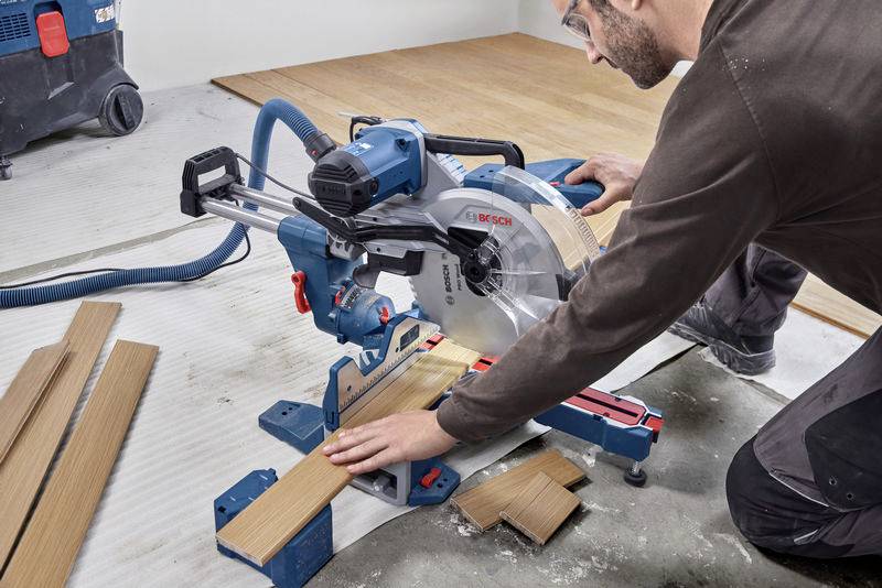 Ein Mann schneidet mit einer blauen Gehrungssäge Holzlatten auf einem Boden. Die Säge ist an einen Staubsauger angeschlossen.
