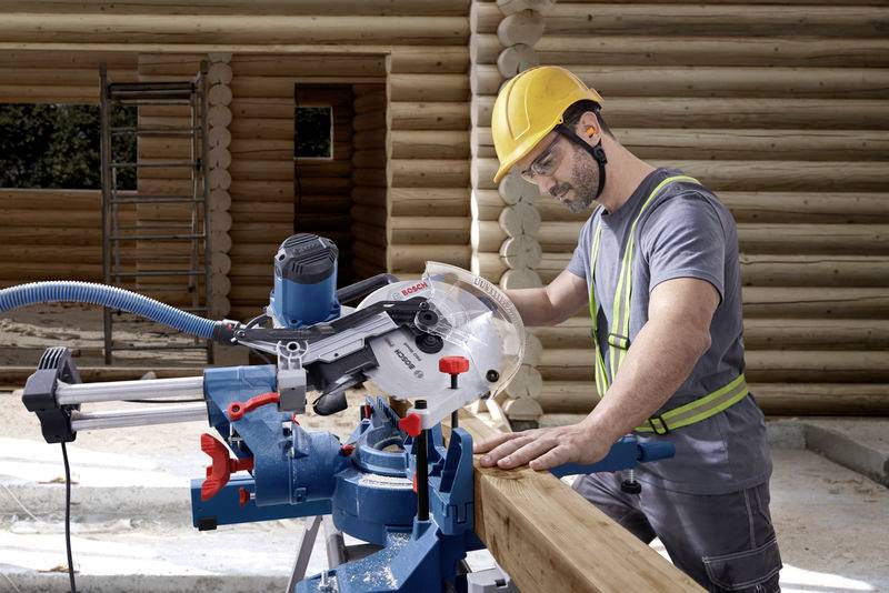 Ein Bauarbeiter mit Schutzhelm und Brille sägt ein Holzbrett auf einer Baustelle. Gebäude aus Holz ist im Hintergrund sichtbar.