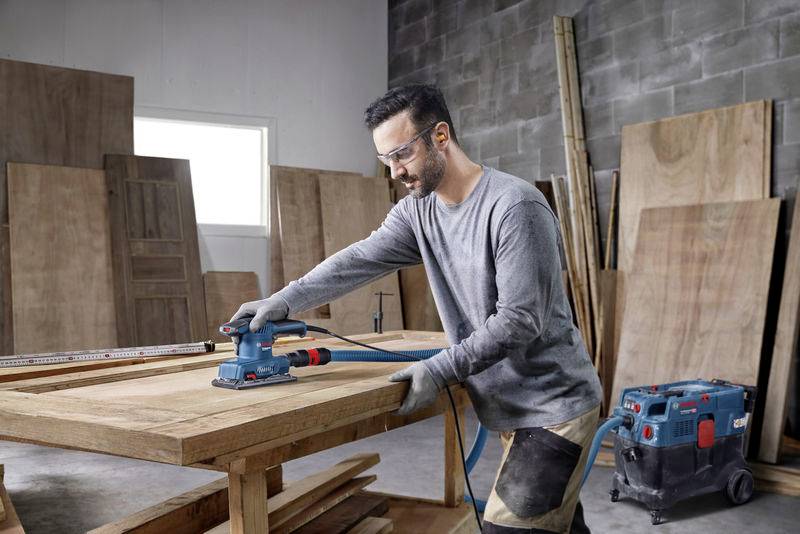 Ein Mann schleift Holz mit einer Handschleifmaschine in einer Werkstatt. Im Hintergrund sind Holzplatten und Werkzeuge sichtbar.