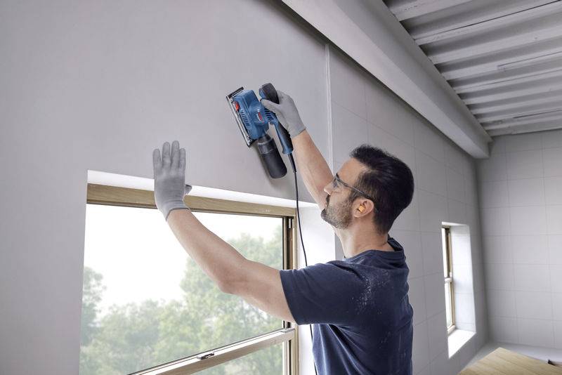 Ein Mann benutzt ein Elektrowerkzeug, um eine Wand nahe einem Fenster zu bearbeiten. Er trägt Schutzbrille und Handschuhe.