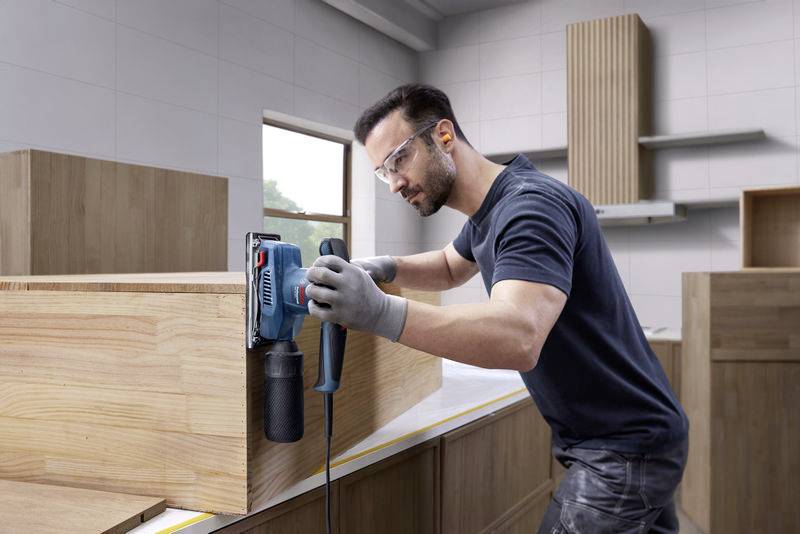 Ein Mann schleift eine Holzoberfläche mit einer elektrischen Schleifmaschine in einer Werkstatt. Er trägt Schutzbrille und Handschuhe.