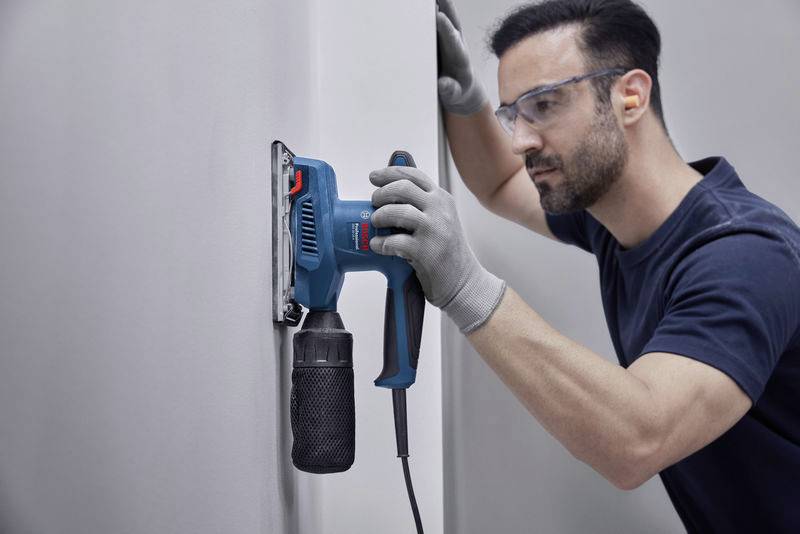 Ein Mann schleift eine Wand mit einem Elektroschleifer. Er trägt Schutzbrille und Handschuhe und arbeitet konzentriert.