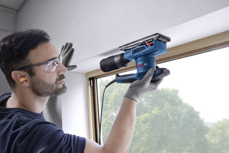 Ein Mann mit Schutzbrille verwendet eine elektrische Schleifmaschine, um ein Fensterbrett zu bearbeiten. Im Hintergrund ist grüne Landschaft sichtbar.
