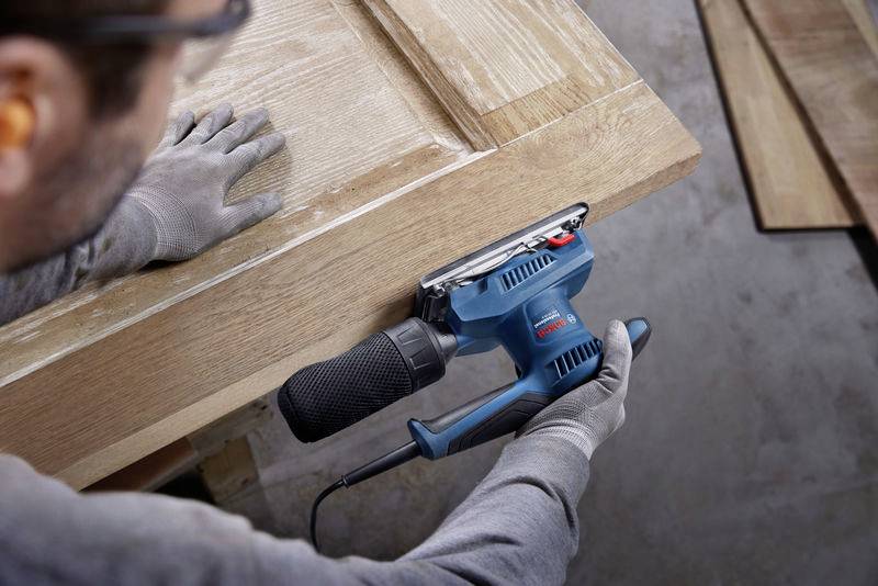 Eine Person schleift mit einem blauen Elektroschleifer eine Holztür, um die Kanten zu glätten. Schutzbrille und Handschuhe sind erkennbar.