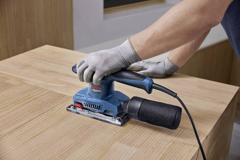 Eine Person schleift eine Holzoberfläche mit einem elektrischen Schleifgerät. Sie trägt Handschuhe für den Schutz.