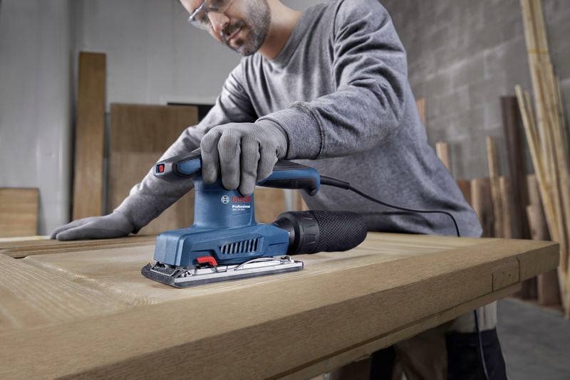 Eine Person schleift mit einem blauen Elektroschleifer eine Holzplatte in einer Werkstatt. Im Hintergrund sind Holzmaterialien zu sehen.