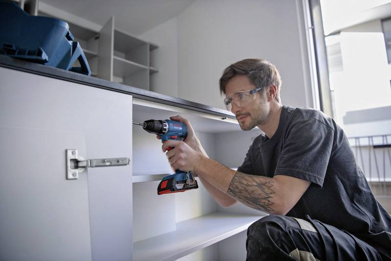 Ein Mann bohrt in ein Scharnier eines Küchenschranks. Er trägt Schutzbrille und arbeitet konzentriert in einer modernen Küche.