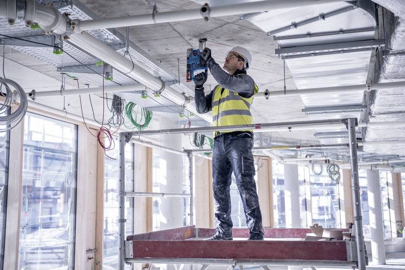 Ein Bauarbeiter in Sicherheitsweste und Helm steht auf einem Gerüst und bohrt Löcher in die Decke eines halbfertigen Gebäudes.