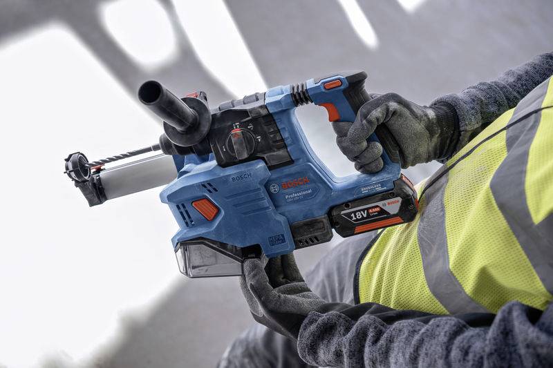 Eine Person hält einen blauen Bosch-Akkubohrhammer mit 18V-Batterie, trägt einen gelben Sicherheitsweste und Arbeitshandschuhe.