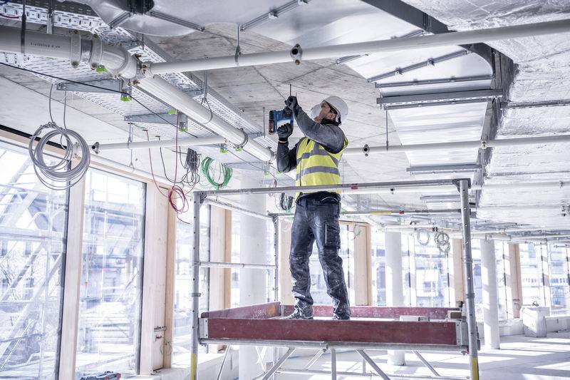 Ein Bauarbeiter steht auf einem Gerüst in einem Gebäude, trägt eine Sicherheitsweste und Helm, und bohrt in die Decke.