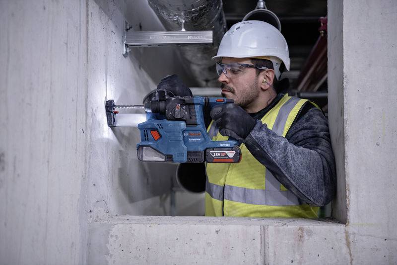 Ein Bauarbeiter trägt einen Schutzhelm und eine Sicherheitsweste, während er mit einem elektrischen Bohrer in eine Betonwand bohrt.