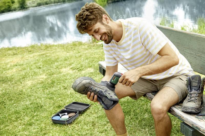 Ein Mann sitzt auf einer Bank am See und reinigt mit einem elektrischen Gerät seine Wanderschuhe. Daneben liegt ein offenes Schuhpflege-Set.