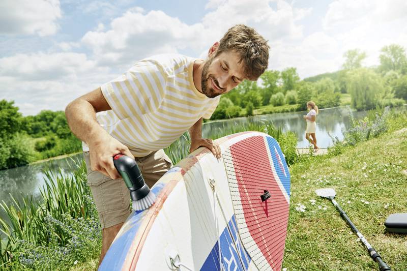 Ein Mann reinigt ein Surfbrett mit einer Bürste an einem Seeufer. Im Hintergrund sieht man eine Person am Wasser stehen.