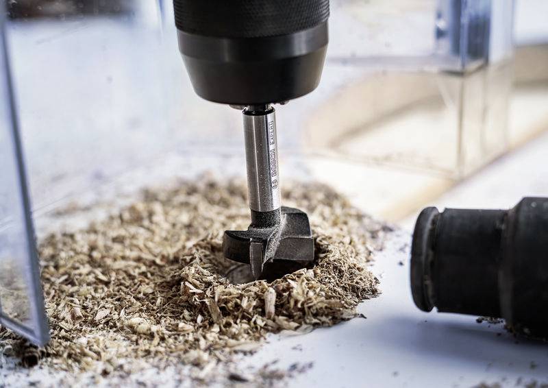 An einem Tisch bohrt ein Bohrer durch Holz, umgeben von Holzspänen. Der Bohrer ist teilweise in das Holz eingedrungen.