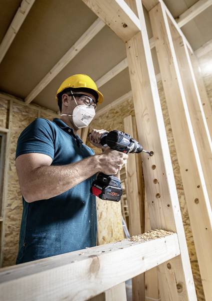 Bauarbeiter trägt Sicherheitsausrüstung und bohrt mit einer elektrischen Bohrmaschine Holzrahmen in einem Rohbau.