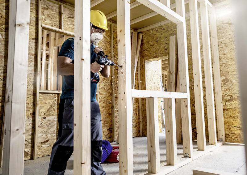Ein Bauarbeiter trägt einen gelben Schutzhelm und eine Maske, während er in einem Holzrahmen-Hausbau mit einer Bohrmaschine arbeitet.