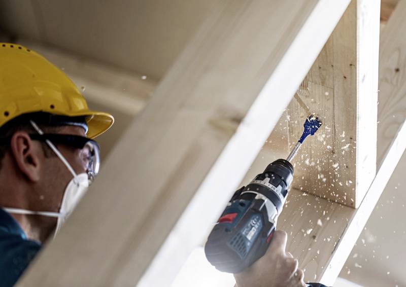 Ein Bauarbeiter trägt Schutzkleidung und benutzt eine elektrische Bohrmaschine, um ein Loch in einen Holzbalken zu bohren. Holzspäne fliegen umher.