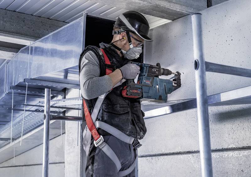Ein Bauarbeiter mit Schutzhelm und Atemmaske bohrt mit einer blauen Elektrowerkzeug an einer Betonwand auf einer Baustelle.