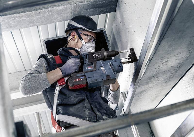 Ein Bauarbeiter bohrt mit einem Elektrowerkzeug in eine Betonwand. Er trägt Schutzkleidung, Helm und Gehörschutz.