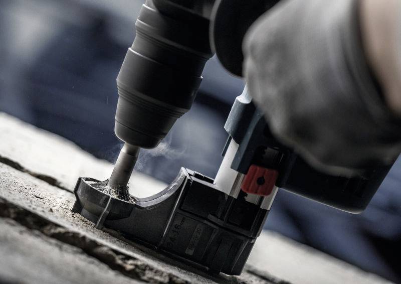 Eine Nahaufnahme eines Bohrers, der in Beton bohrt, mit Staub, der von der Bohrstelle aufsteigt. Eine behandschuhte Hand hält den Bohrer fest.