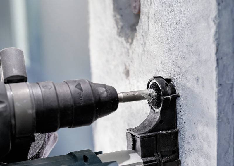 Ein Bohrer bricht in eine Betonwand ein, während er von einer Person bedient wird. Der Fokus liegt auf dem Bohren in den Beton.