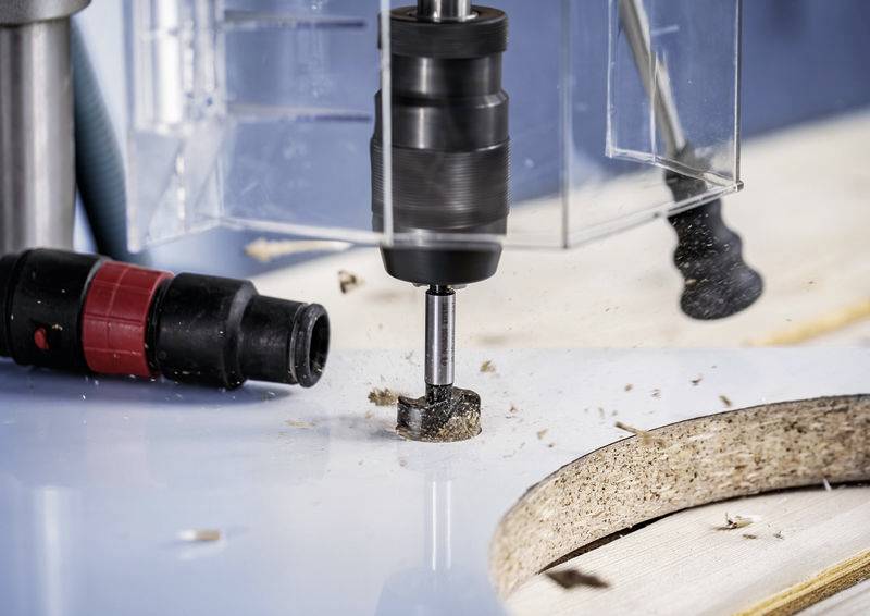 Ein Bohrer wird verwendet, um ein rundes Loch in ein Holzbrett zu bohren. Holzspäne sind auf der Arbeitsfläche sichtbar.