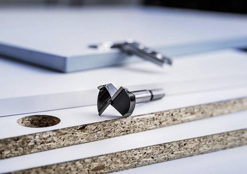 Ein Bohrkopf und Schichten von Spanplatten mit einem runden Loch. Zeigt Werkzeuge und Material für Holzarbeiten.