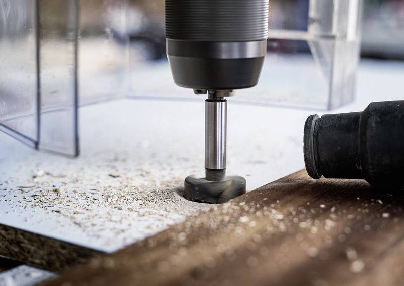 Eine Bohrmaschine bearbeitet Holz, mit Holzspänen und Staub auf der Oberfläche. Die Maschine ist auf ein Stück Holz ausgerichtet.