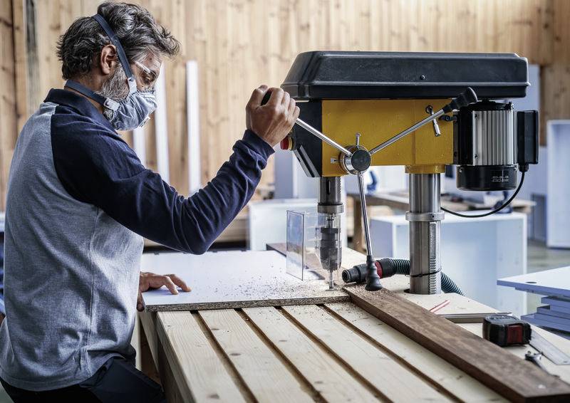 Ein Mann mit Schutzbrille und Maske arbeitet an einer Bohrmaschine in einer Werkstatt. Holzspäne fallen auf den Tisch.
