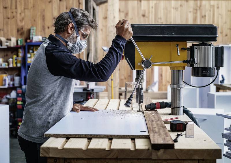 Eine Person mit Schutzmaske arbeitet an einer Standbohrmaschine in einer Werkstatt. Im Hintergrund sind Holzregale und Werkzeuge sichtbar.