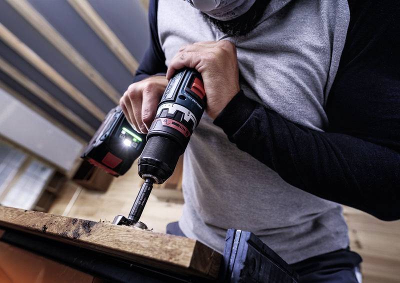 Person arbeitet mit einer Bohrmaschine an einem Holzstück in einer Werkstattumgebung.