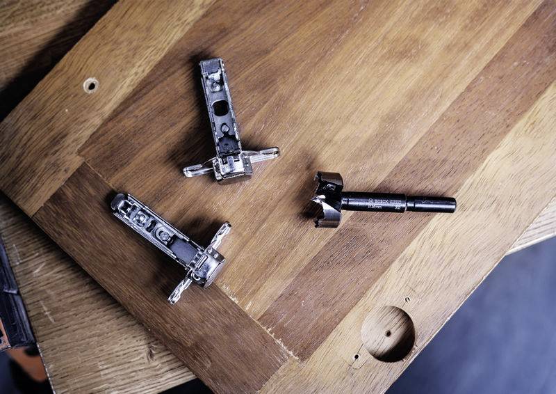 Drei Scharniere und ein Bohrkopf liegen auf einem Holztisch. Die Komponenten sind vermutlich für den Zusammenbau von Möbeln gedacht.