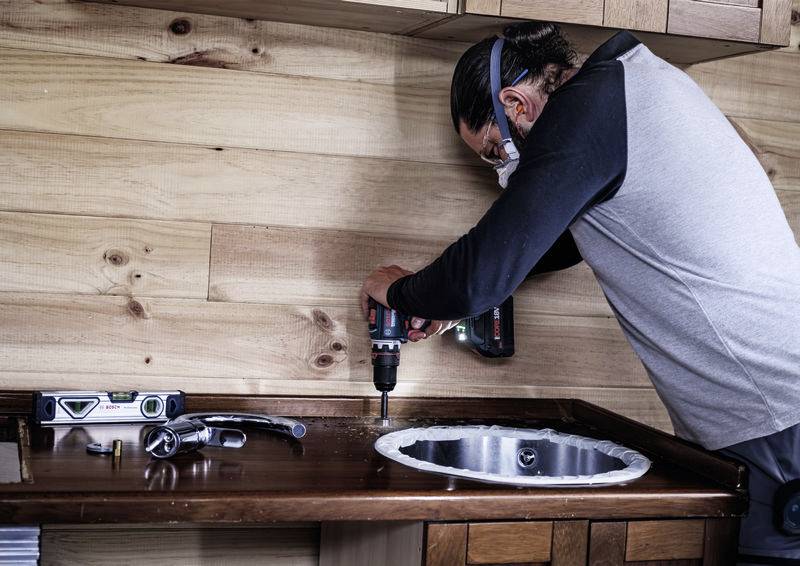 Handwerker installiert eine Küchenspüle, benutzt einen Akkuschrauber. Mess- und Handwerkzeug liegen daneben auf der Arbeitsplatte.
