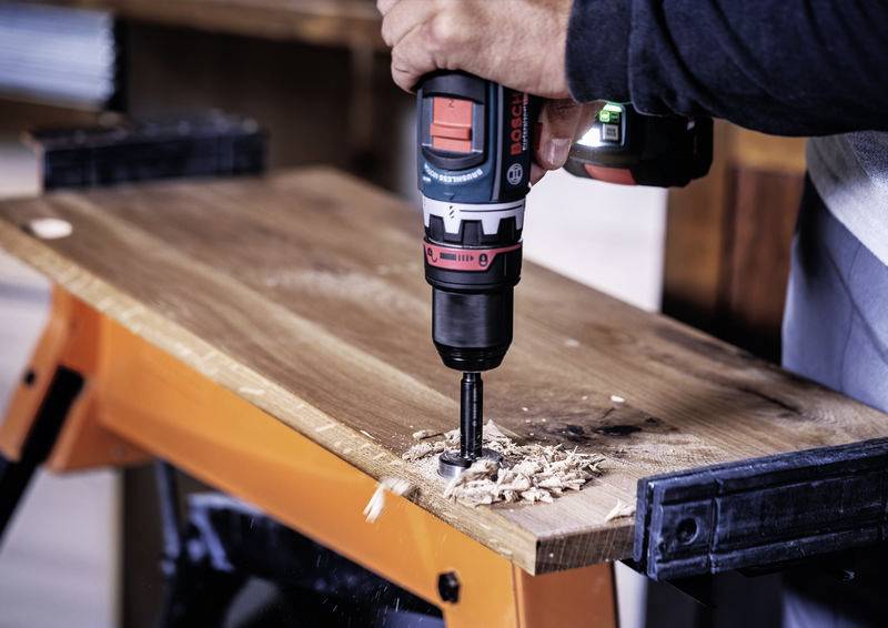 Ein Mensch bohrt mit einer elektrischen Bohrmaschine ein Loch in ein Holzbrett, das auf einer Werkbank liegt. Holzspäne sind sichtbar.