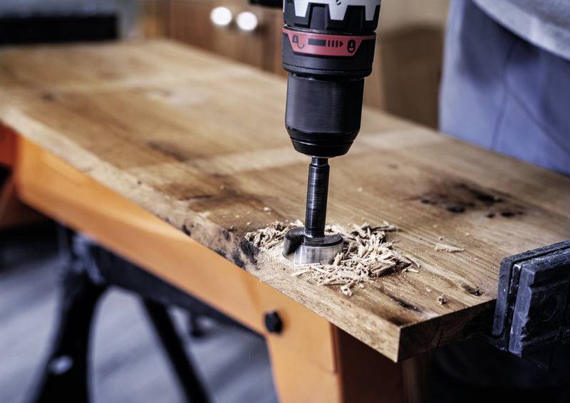 Person verwendet eine Bohrmaschine, um ein Loch in ein Holzbrett zu bohren. Holzspäne sind um das Bohrloch verteilt.