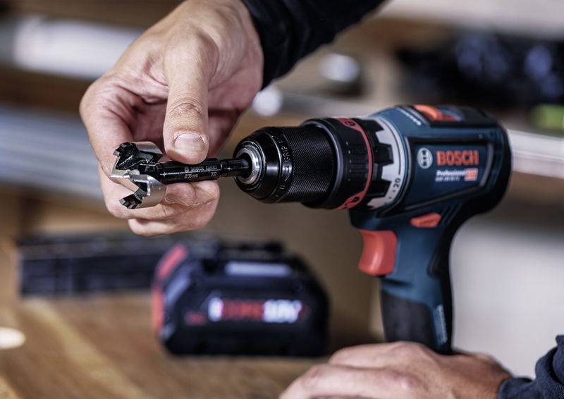 Eine Hand hält einen Bohrer, der in einen elektrischen Akkuschrauber eingesetzt wird. Im Hintergrund ist unscharf eine Akku-Box erkennbar.