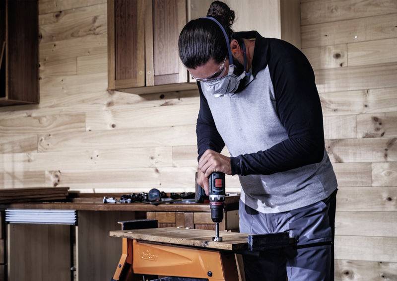 Eine Person arbeitet in einer Holzwerkstatt und bohrt ein Stück Holz auf einer Werkbank. Sie trägt Schutzbrille und Maske.