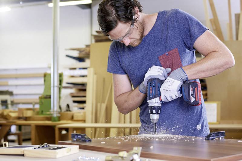 Ein Mann in einer Werkstatt bohrt präzise mit einer elektrischen Bohrmaschine in ein Holzstück und trägt dabei Schutzbrille und Handschuhe.