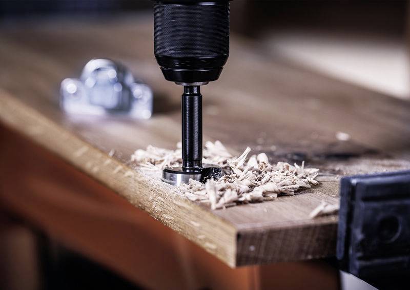 Eine Bohrmaschine fräst ein Loch in ein Holzbrett, Holzspäne liegen daneben. Sie ist an der Kante eines Werkstatttisches befestigt.