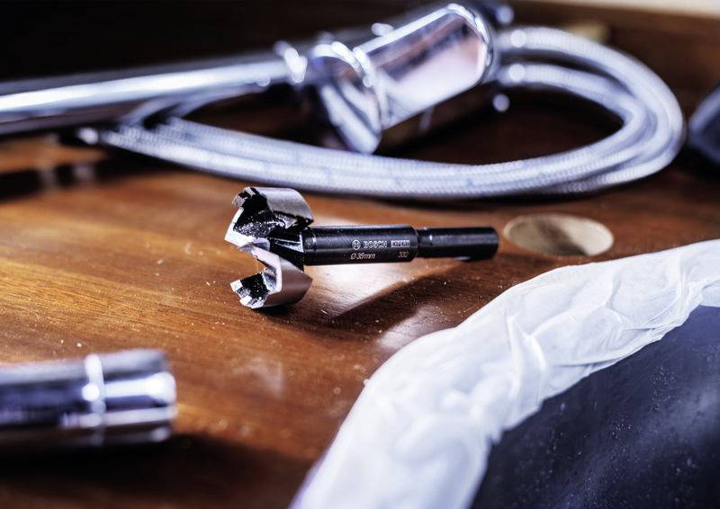 Nahaufnahme eines 28-mm-Bohrers auf einem Holztisch, umgeben von einem flexiblen Metallrohr und einem verchromten Werkzeug.