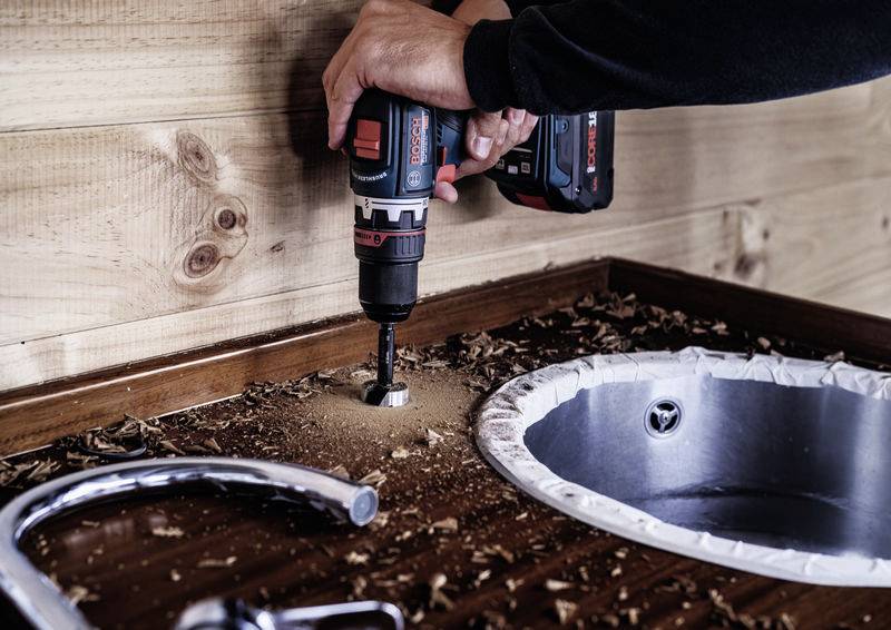 Eine Person bohrt mit einer Bohrmaschine ein Loch in eine Holzplatte neben einem Spülbecken. Holzspäne sind verstreut.