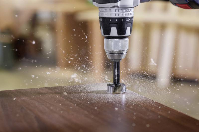 Ein Forstnerbohrer wird verwendet, um präzise Löcher in ein Holzbrett zu bohren. Bohrspäne fliegen in die Luft.