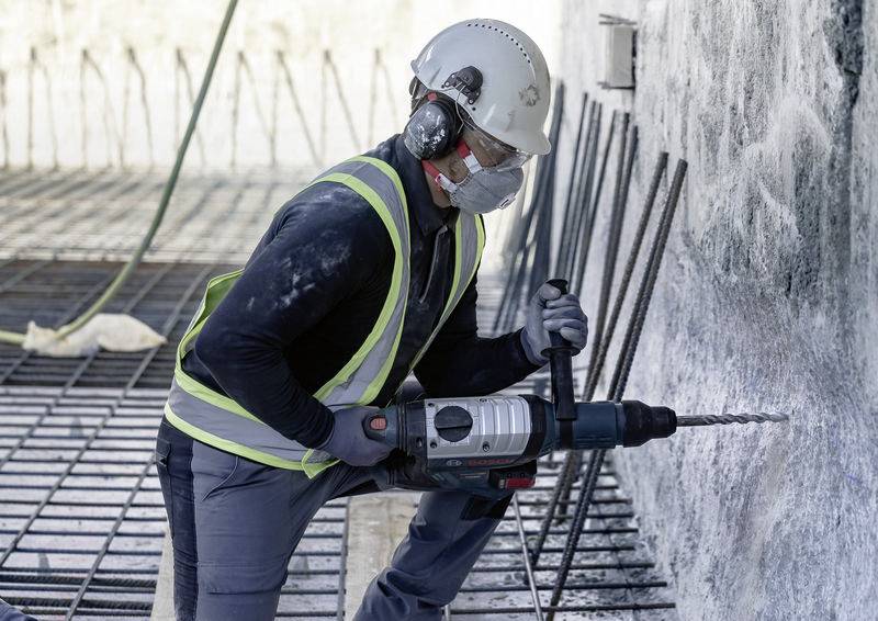 Arbeiter mit Schutzkleidung benutzt eine Bohrmaschine an einer Betonwand auf einer Baustelle. Metallelemente im Hintergrund.