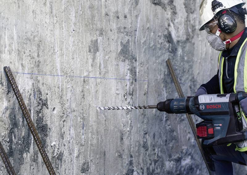 Ein Bauarbeiter in Sicherheitskleidung bohrt mit einer Bohrmaschine ein Loch in eine Betonwand. Eisenstangen ragen aus der Wand.