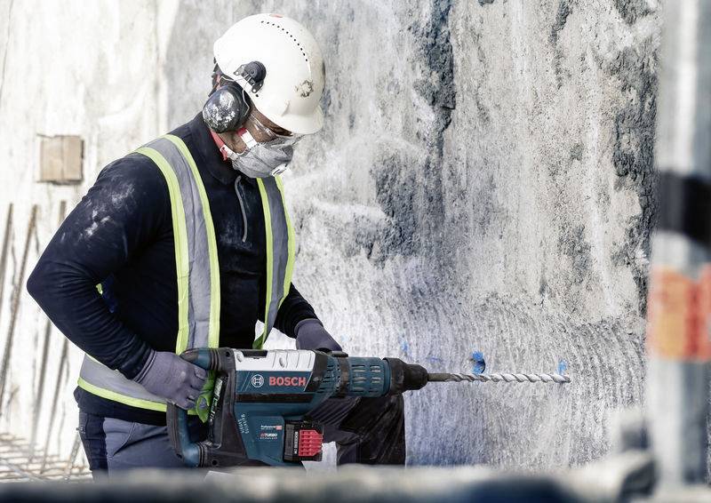 Arbeiter trägt Schutzausrüstung und bohrt mit einer Bosch-Maschine in eine Betonwand, Baustellenhintergrund deutlich erkennbar.